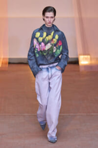 Model walking down a runway in a navy floral-print shirt and oversized light jeans, hands in pockets.