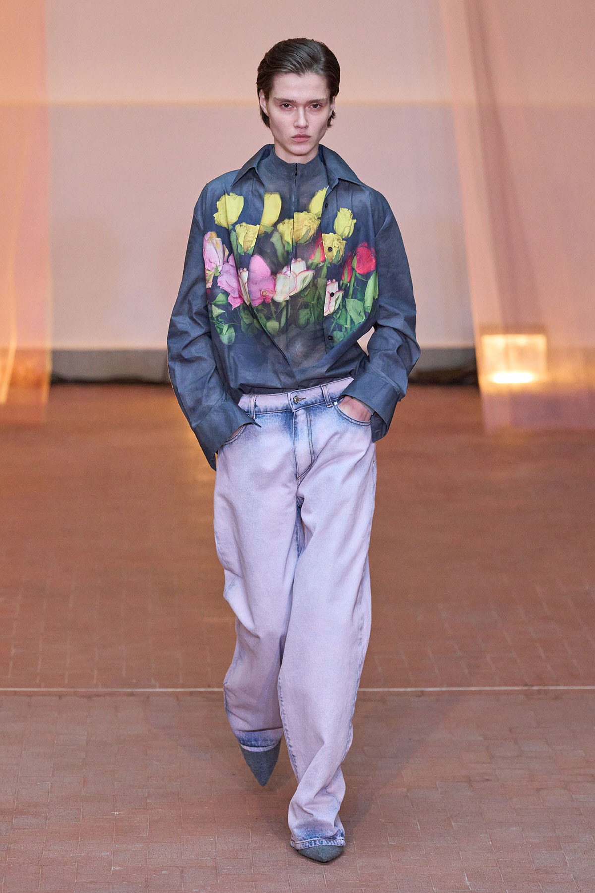 Model walking down a runway in a navy floral-print shirt and oversized light jeans, hands in pockets.
