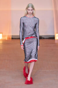 Model walking a runway in a long-sleeve gray marble-pattern dress with red and white trim, paired with bright red fluffy slippers.
