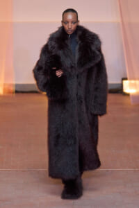 Model walks the runway wearing an oversized black fur coat with sleek hair and neutral makeup in a fashion show setting.