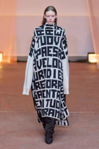 Runway model wearing an oversized black-and-white geometric poncho with bold letter blocks, white-sleeved top underneath, and black pants with boots.