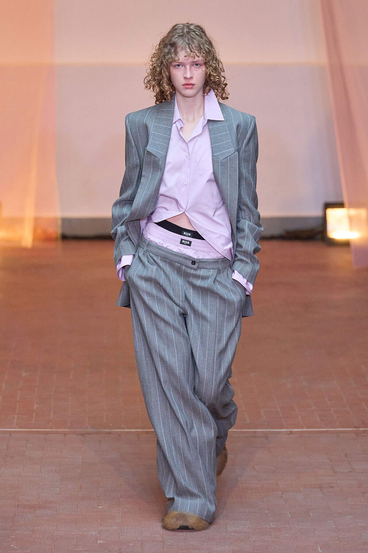 Model walking a runway in an oversized gray pinstripe suit with a tucked pink shirt, hands in pockets, curly hair, showing a relaxed yet edgy look.