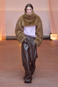 Model walking a runway in a voluminous brown fur coat over a lavender blouse and dark brown leather wide-leg pants.