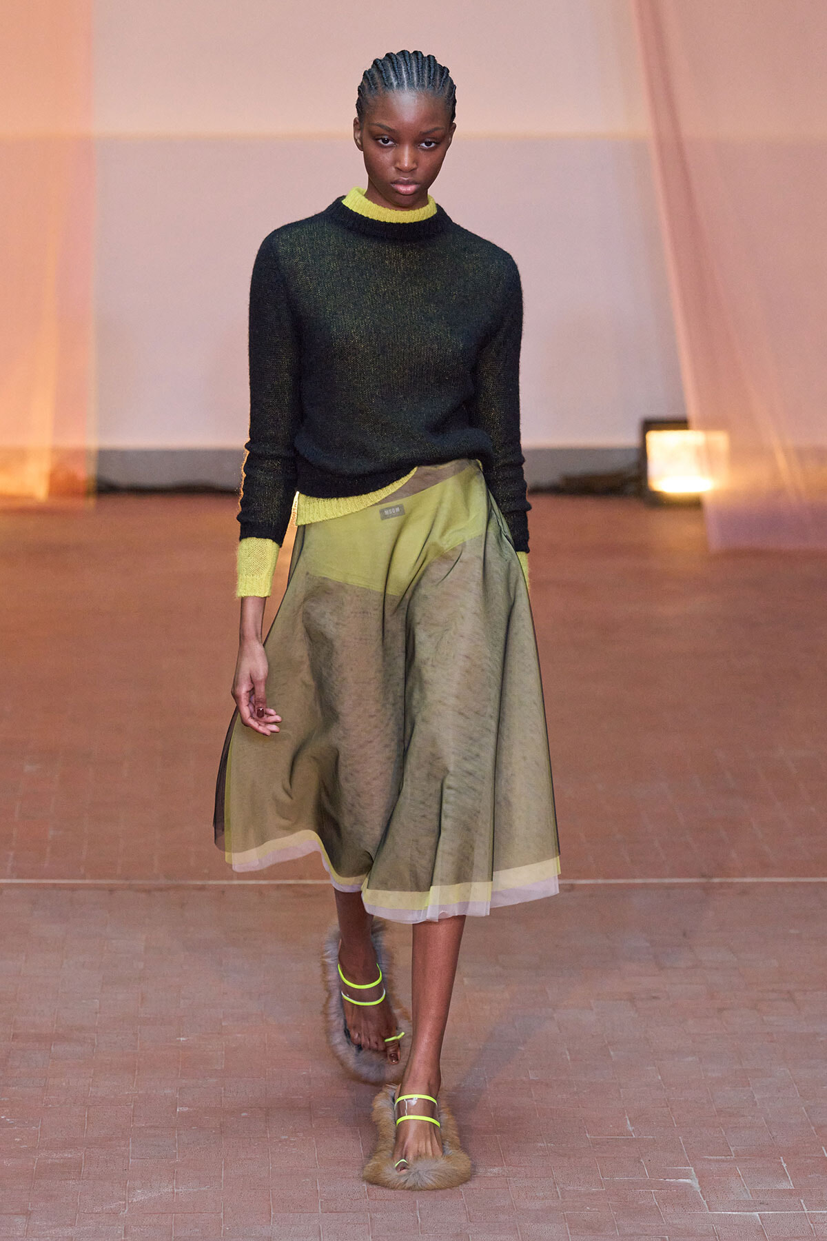 Model walking a runway in a black knit sweater with lime cuffs and collar, an olive midi skirt, and neon-green fur-trimmed sandals.