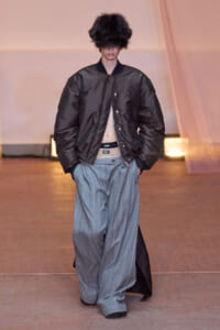 Model on a runway wearing a black oversized bomber jacket, wide gray pinstripe trousers, and a large black fur hat obscuring the eyes.