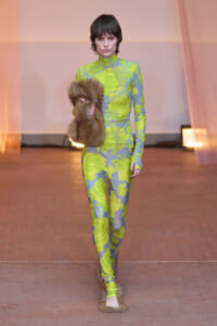 Model on runway wearing neon lime-green camouflage bodysuit, holding a brown fur stole.
