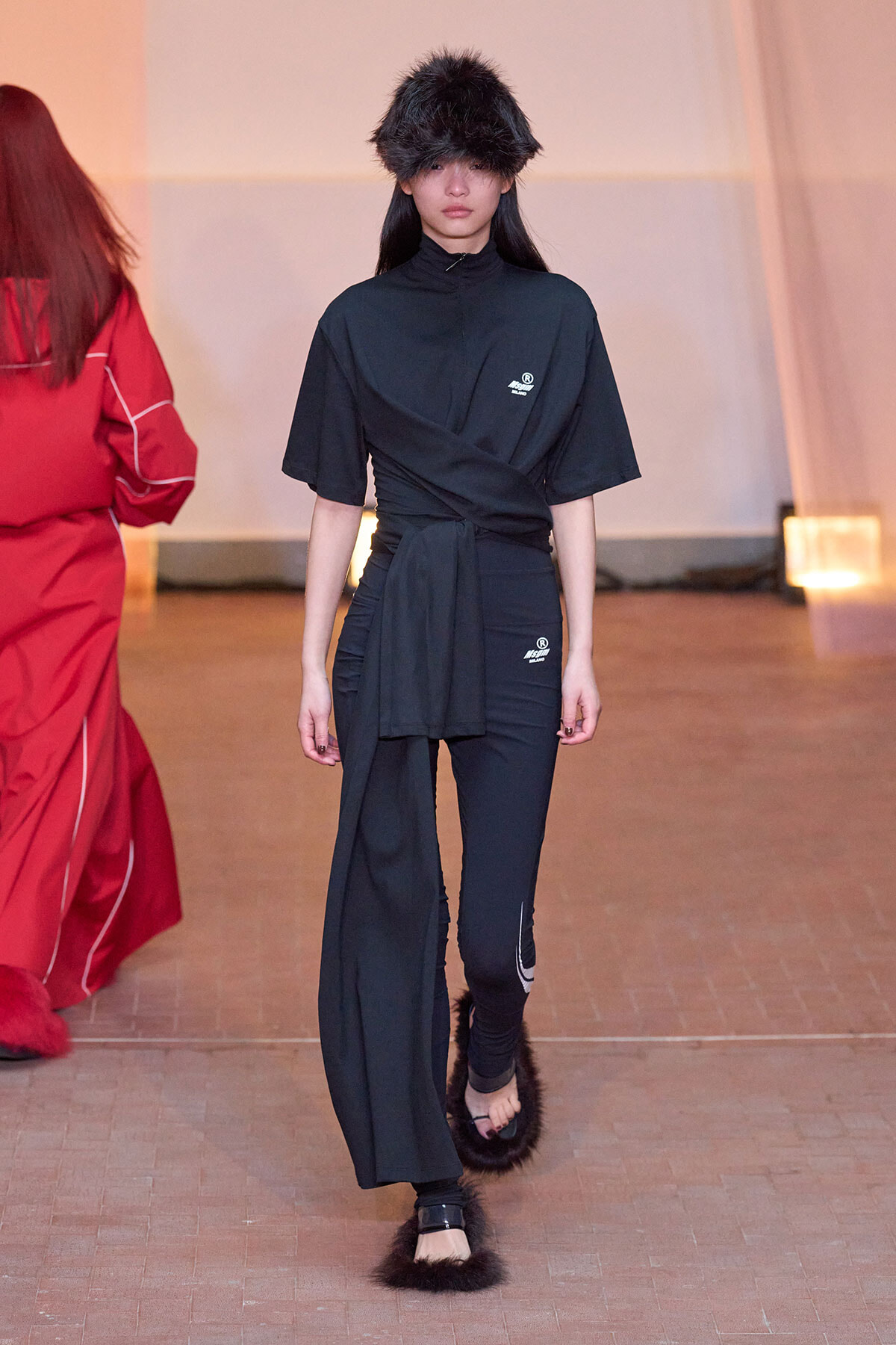 Model walks the runway in a black wrap-top outfit with matching pants and fur hat, minimalist staging in warm lighting.