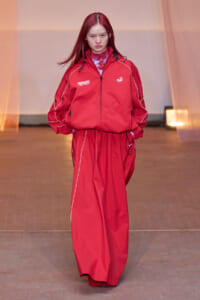 Female model walking a fashion show in a red windbreaker and matching wide-leg pants with white piping.
