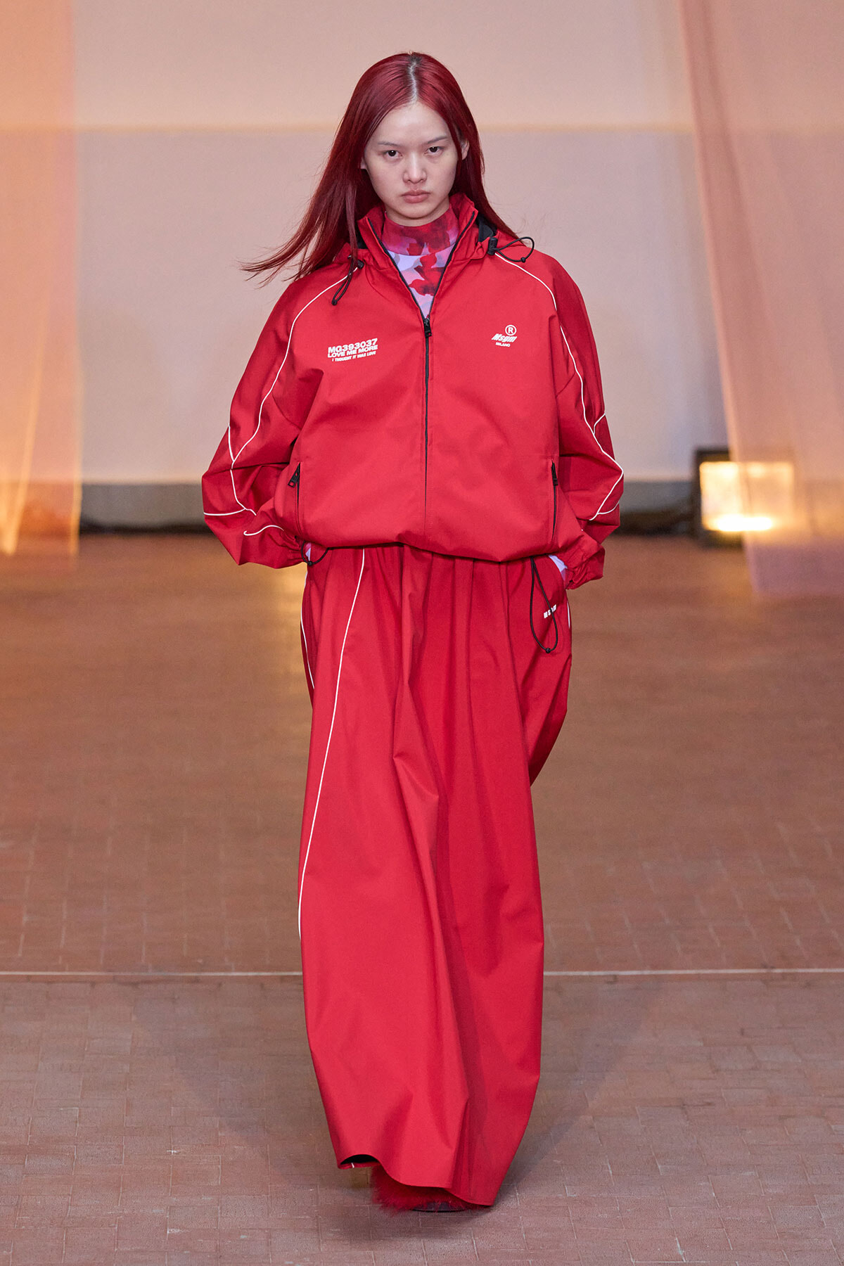 Female model walking a fashion show in a red windbreaker and matching wide-leg pants with white piping.