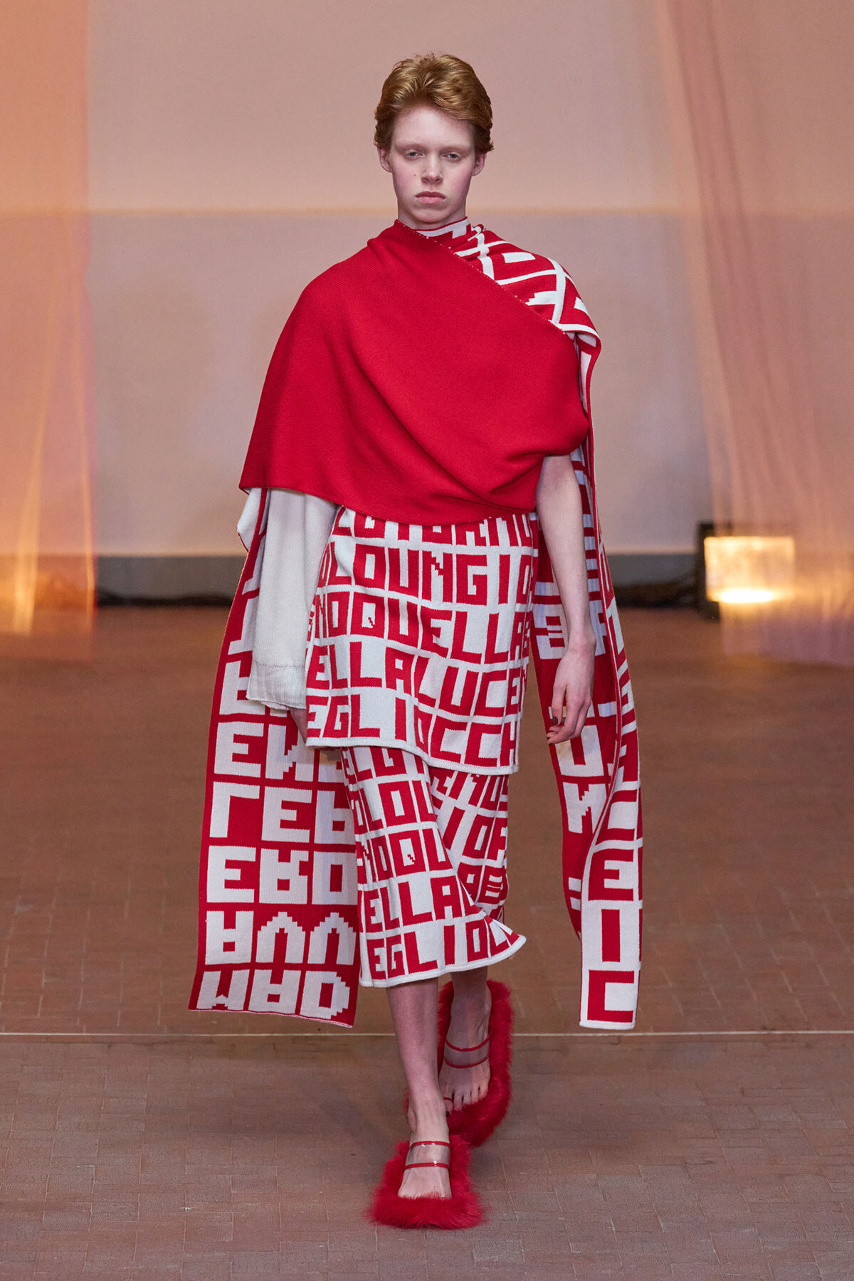 Model walks the runway in a bold red cape over a white-and-red graphic outfit with large block letters; red fluffy shoes complete the look.