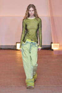 Runway model walks the catwalk in a sheer olive-green top and oversized light-green jeans with yellow accents and trim.