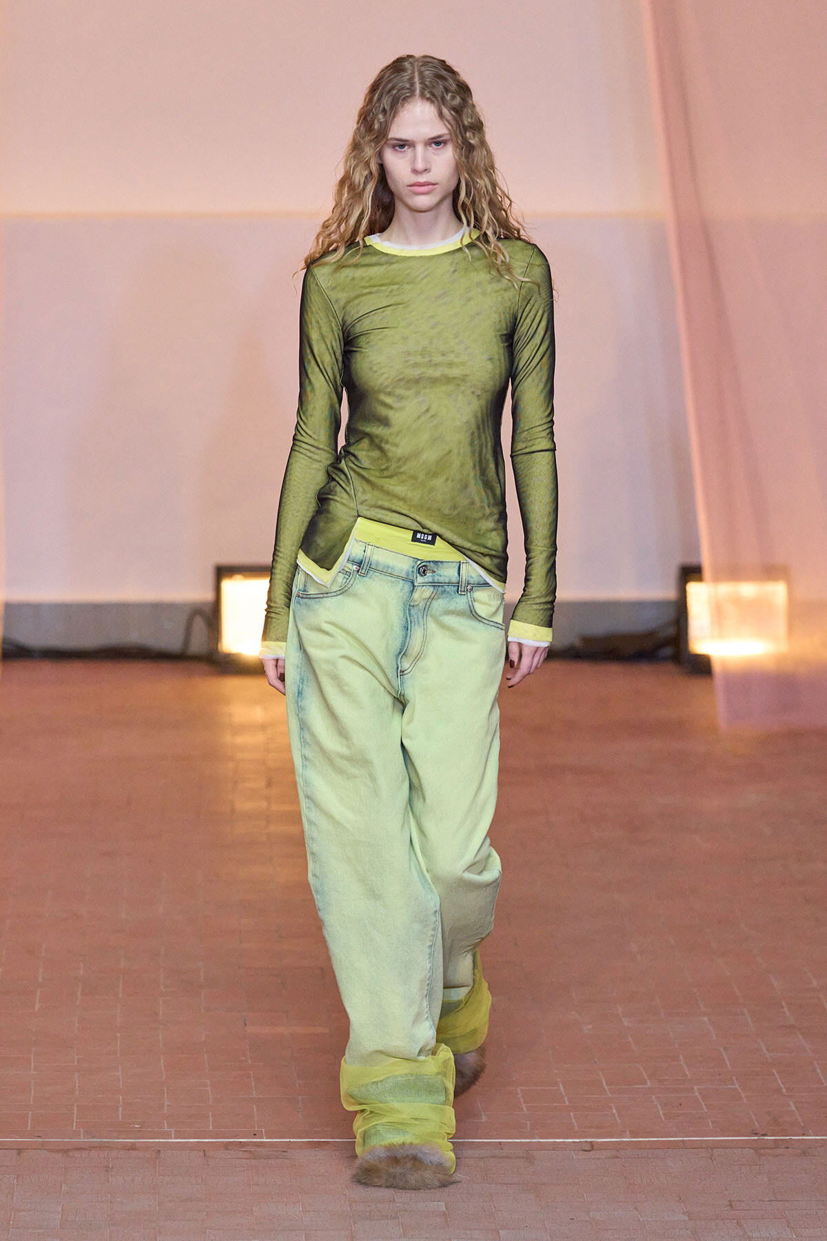 Runway model walks the catwalk in a sheer olive-green top and oversized light-green jeans with yellow accents and trim.