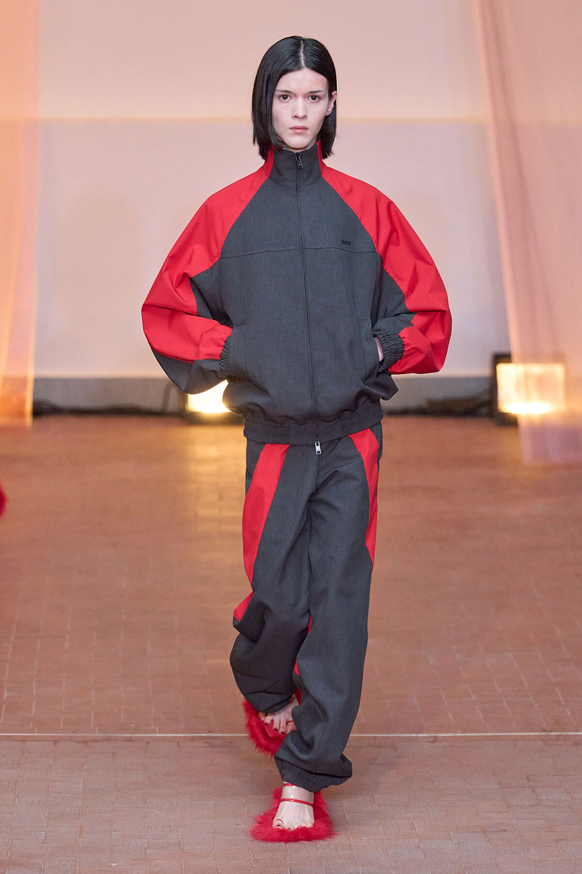Model walking a fashion show wearing a grey and red color-block track suit with zip jacket and elastic cuffs, red panels on sleeves and legs, and red fluffy slippers.