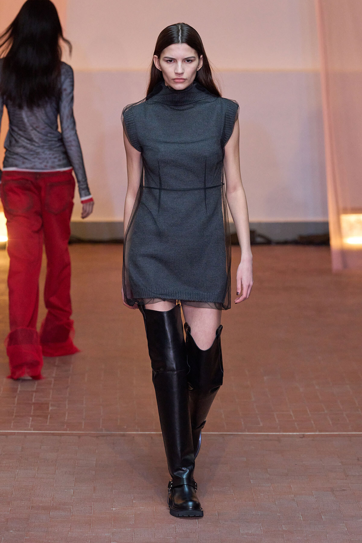 Model walks the runway in a dark gray sleeveless turtleneck dress with sheer side panels, paired with black over‑the‑knee boots at a fashion show.