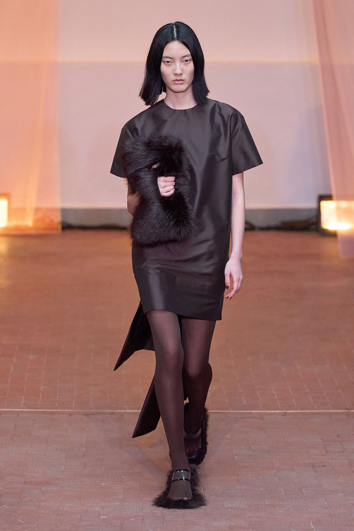 Model walks the runway in a sleek black dress, holding a black fur accessory against a warm, staged backdrop.
