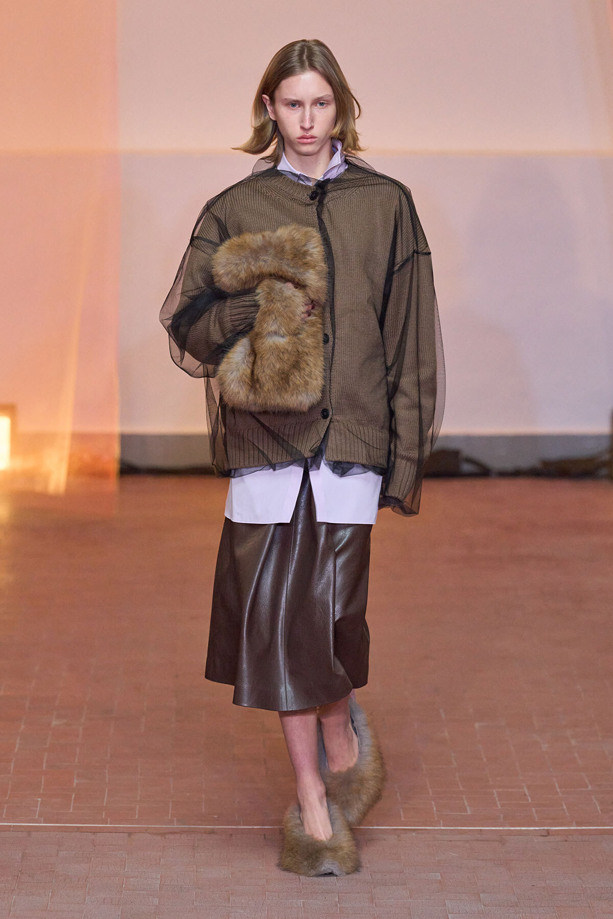 Model on a runway wearing a sheer olive-brown mesh jacket over a light shirt, paired with a brown leather midi skirt and tan fur accessories.