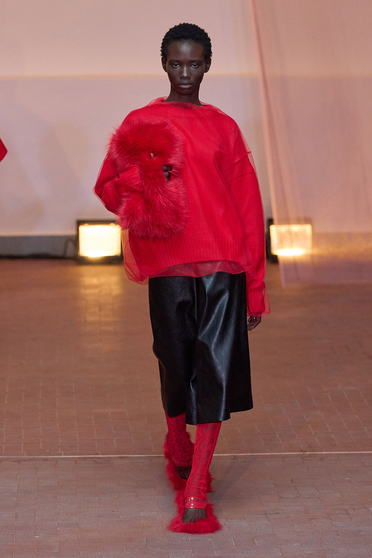 Model on a runway wearing an oversized red knit sweater with a red fur accessory, a black midi skirt, and red lace socks with fur details — bold monochrome look.