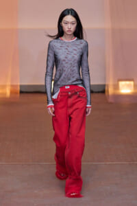 Model walking the runway in a sheer gray long-sleeve top with red trim, paired with oversized red pants and red furry footwear on a warm-toned backdrop.