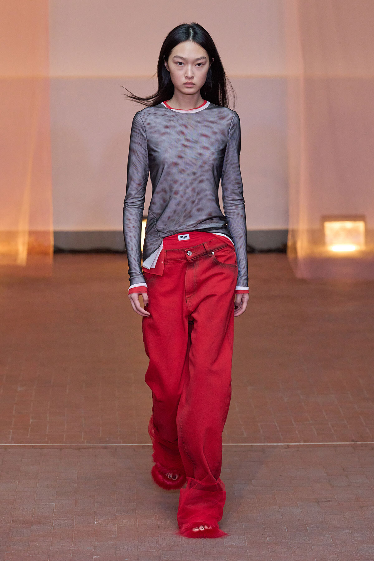 Model walking the runway in a sheer gray long-sleeve top with red trim, paired with oversized red pants and red furry footwear on a warm-toned backdrop.