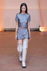 Model walking a runway in a metallic gray two-piece outfit (crop top and shorts) with tall white boots.