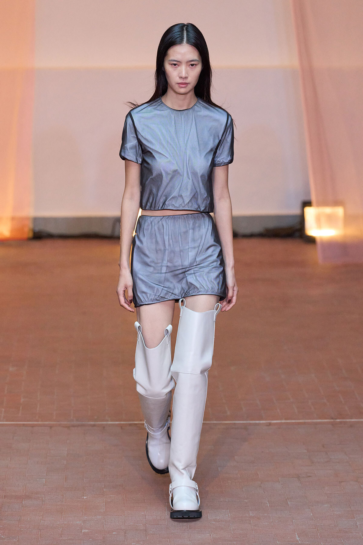 Model walking a runway in a metallic gray two-piece outfit (crop top and shorts) with tall white boots.