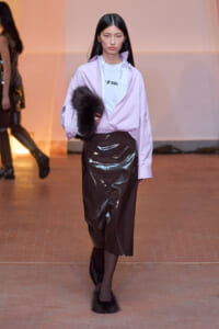 Model walking the runway in a lavender oversized shirt over a white graphic tee, a glossy black vinyl midi skirt, black tights, and fluffy fur-trimmed boots, holding a fur muff.