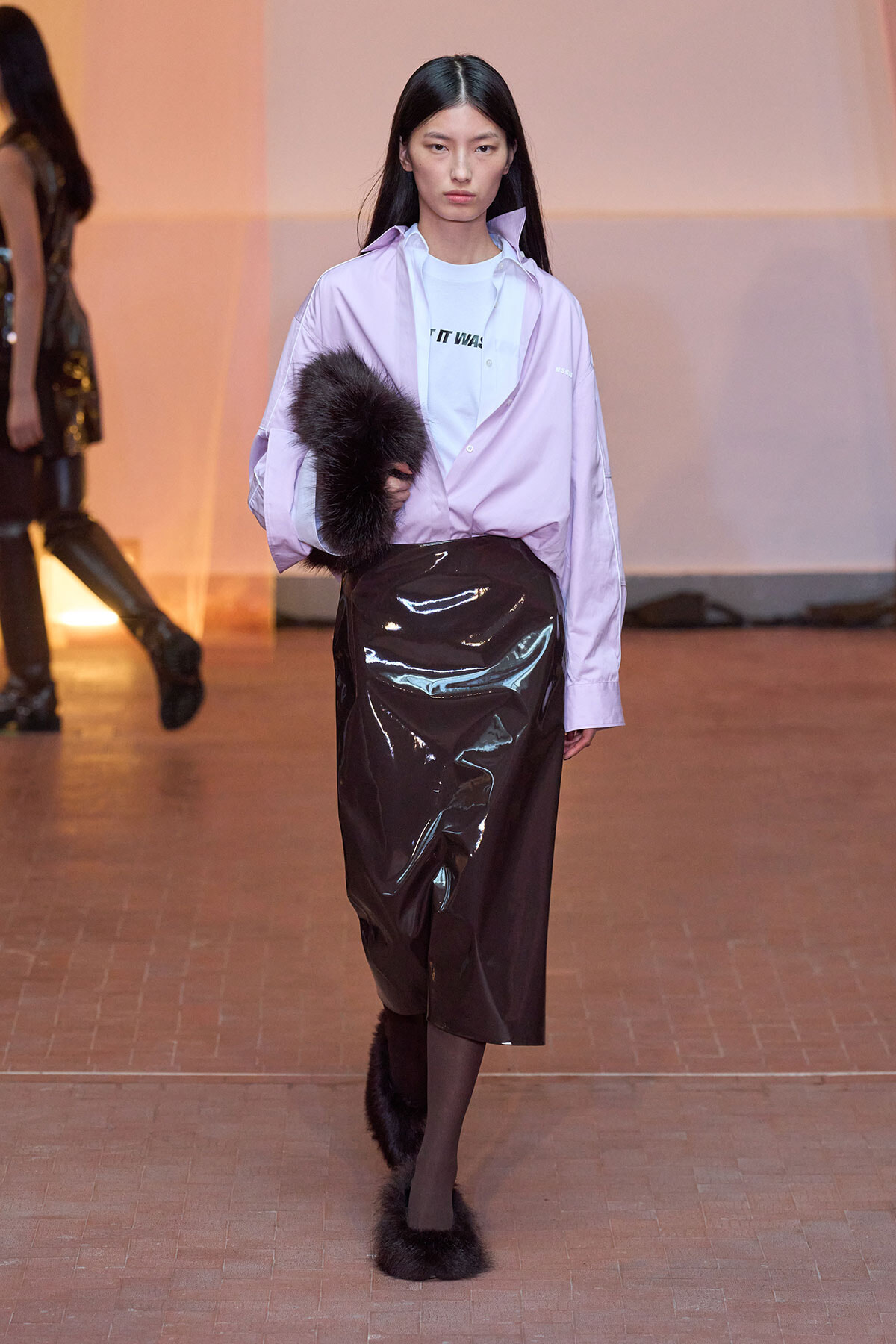Model walking the runway in a lavender oversized shirt over a white graphic tee, a glossy black vinyl midi skirt, black tights, and fluffy fur-trimmed boots, holding a fur muff.