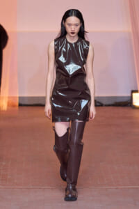 Model walks the runway in a shiny black vinyl sleeveless mini dress and tall brown knee-high boots on a fashion catwalk.