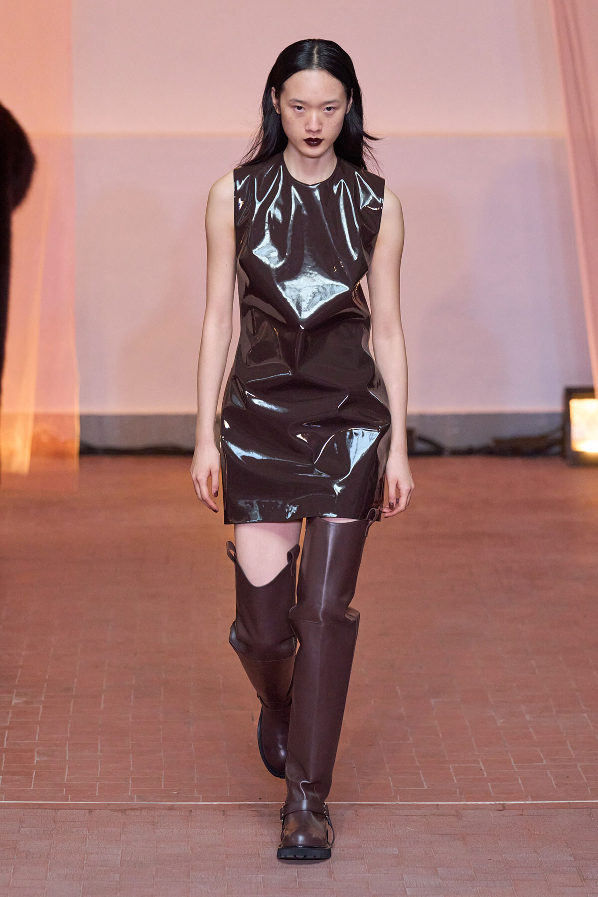 Model walks the runway in a shiny black vinyl sleeveless mini dress and tall brown knee-high boots on a fashion catwalk.