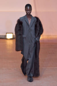 Model walks the runway in a loose gray suit paired with a long dark fur coat, in a warm-lit setting
