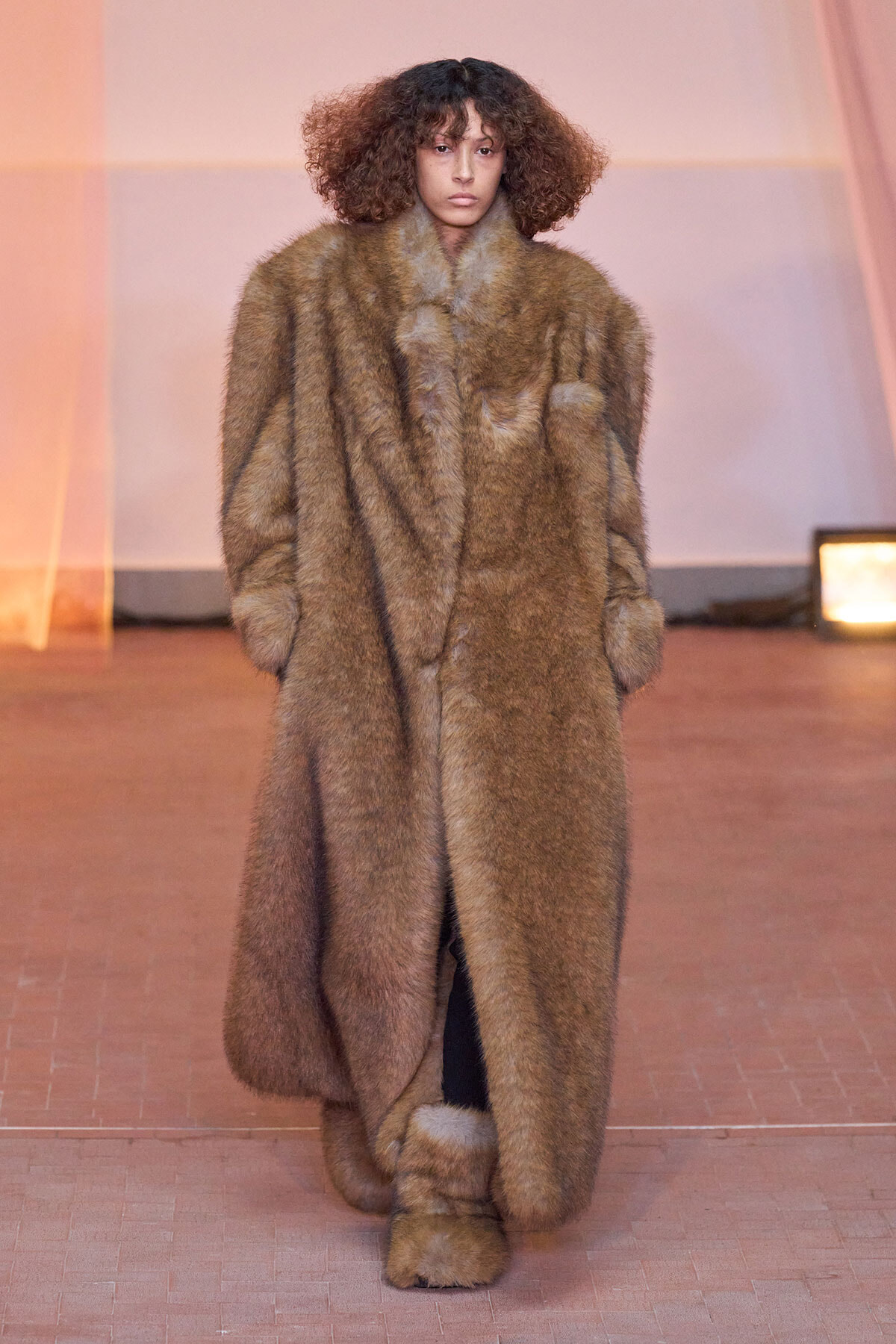 Model walking a fashion runway in a full-length brown fur coat with matching fur boots, neutral expression, soft pink backdrop behind.