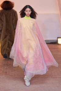 Female model walks the runway wearing a sheer pink cape over a pale-yellow garment, with a neon lime collar peeking out and white shoes.