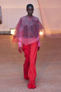 Model walking a runway in a pink sheer cape over a red jumpsuit, pointed red fur-lined shoes.