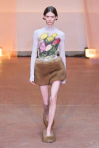 Model walks the runway in a pale floral-printed top paired with a brown fur mini skirt and matching fur footwear amid warm lighting.