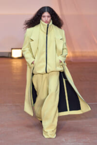 Model walking the runway in a pale yellow oversized coat with black interior panels and zipper, hands in pockets.