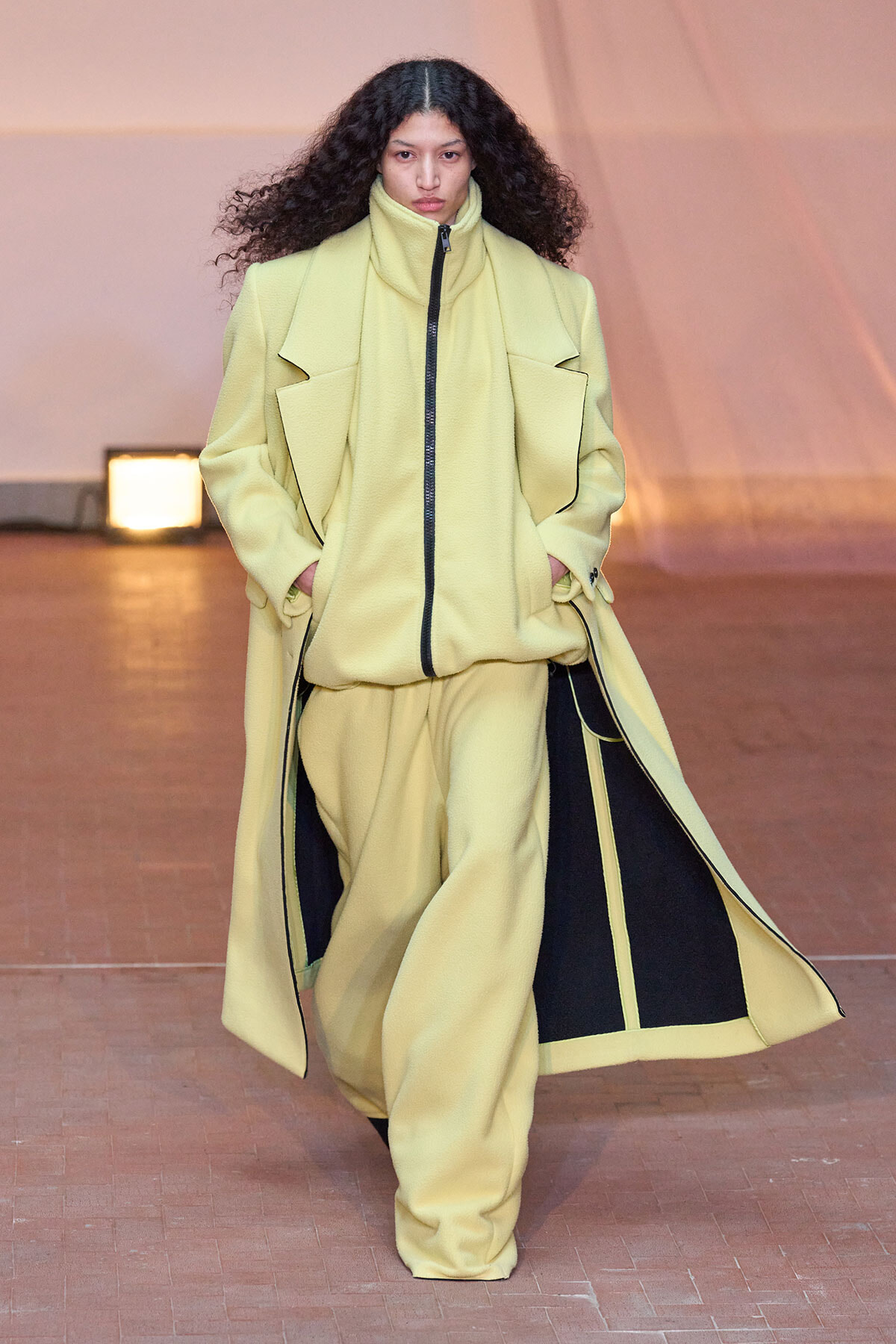 Model walking the runway in a pale yellow oversized coat with black interior panels and zipper, hands in pockets.