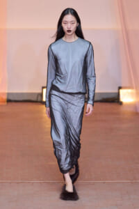 Model on a runway wearing a silver-gray satin long-sleeve top and matching draped pants, paired with fluffy fur slippers and dark lipstick.
