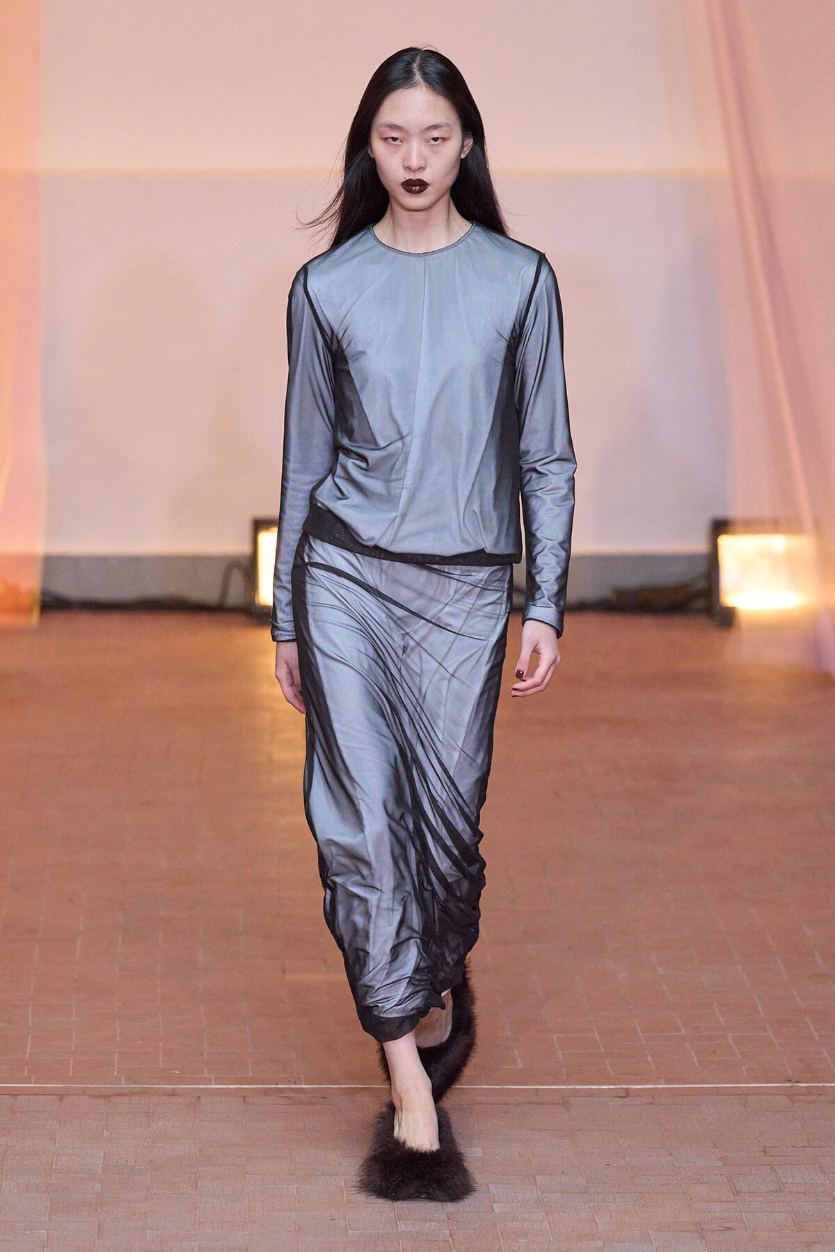 Model on a runway wearing a silver-gray satin long-sleeve top and matching draped pants, paired with fluffy fur slippers and dark lipstick.