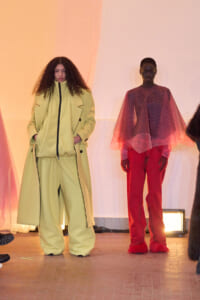 Two fashion models stand on a runway wearing bold color outfits: left in a pale yellow oversized coat and wide-leg pants; right in red pants with a sheer pink cape top.