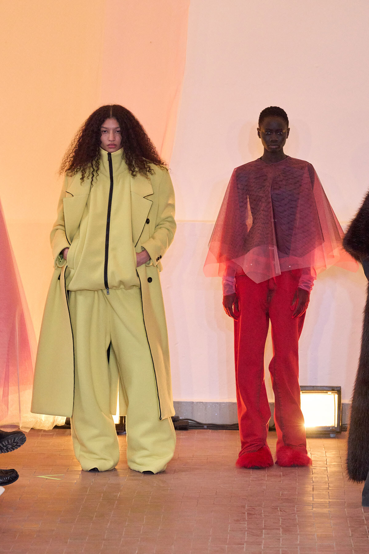 Two fashion models stand on a runway wearing bold color outfits: left in a pale yellow oversized coat and wide-leg pants; right in red pants with a sheer pink cape top.