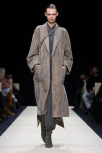 Model on a fashion runway wearing a long gray herringbone coat over a gray outfit with green knee-high boots in a minimalist setting.