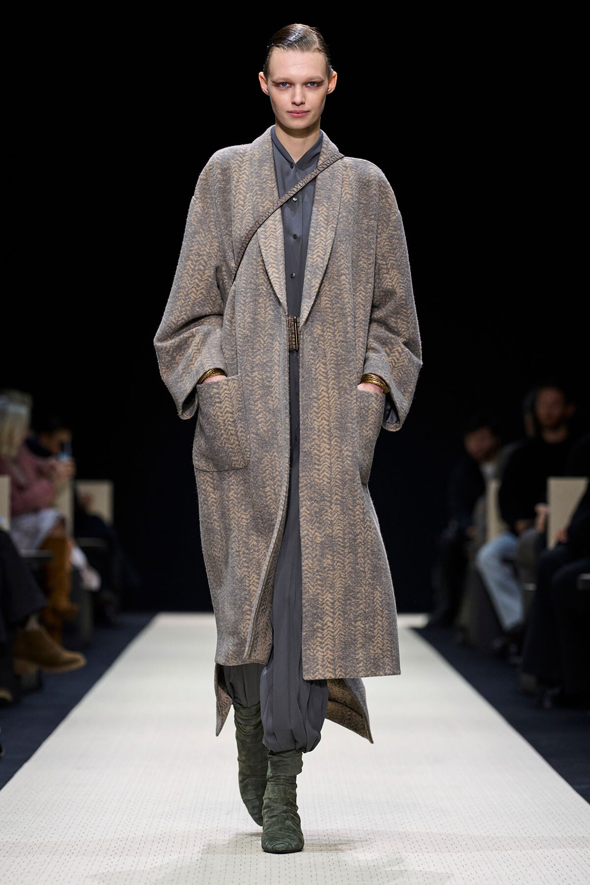 Model on a fashion runway wearing a long gray herringbone coat over a gray outfit with green knee-high boots in a minimalist setting.