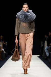 Runway model wearing an oversized gray fur scarf with a brown textured top, peach satin wide-leg pants, brown boots, and a brown handbag.