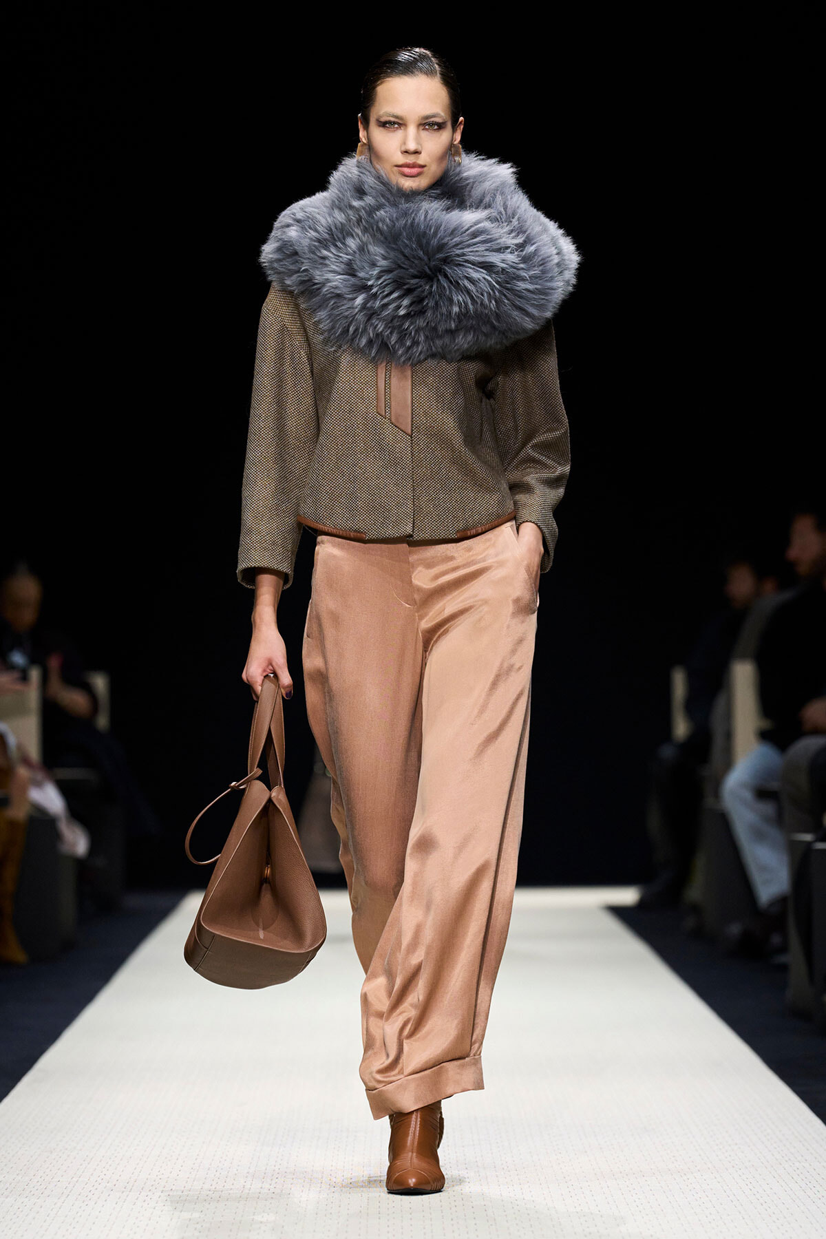 Runway model wearing an oversized gray fur scarf with a brown textured top, peach satin wide-leg pants, brown boots, and a brown handbag.