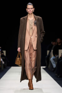 Model walking a runway in a long brown overcoat over a blush-pink suit, carrying a brown handbag and wearing tan boots.
