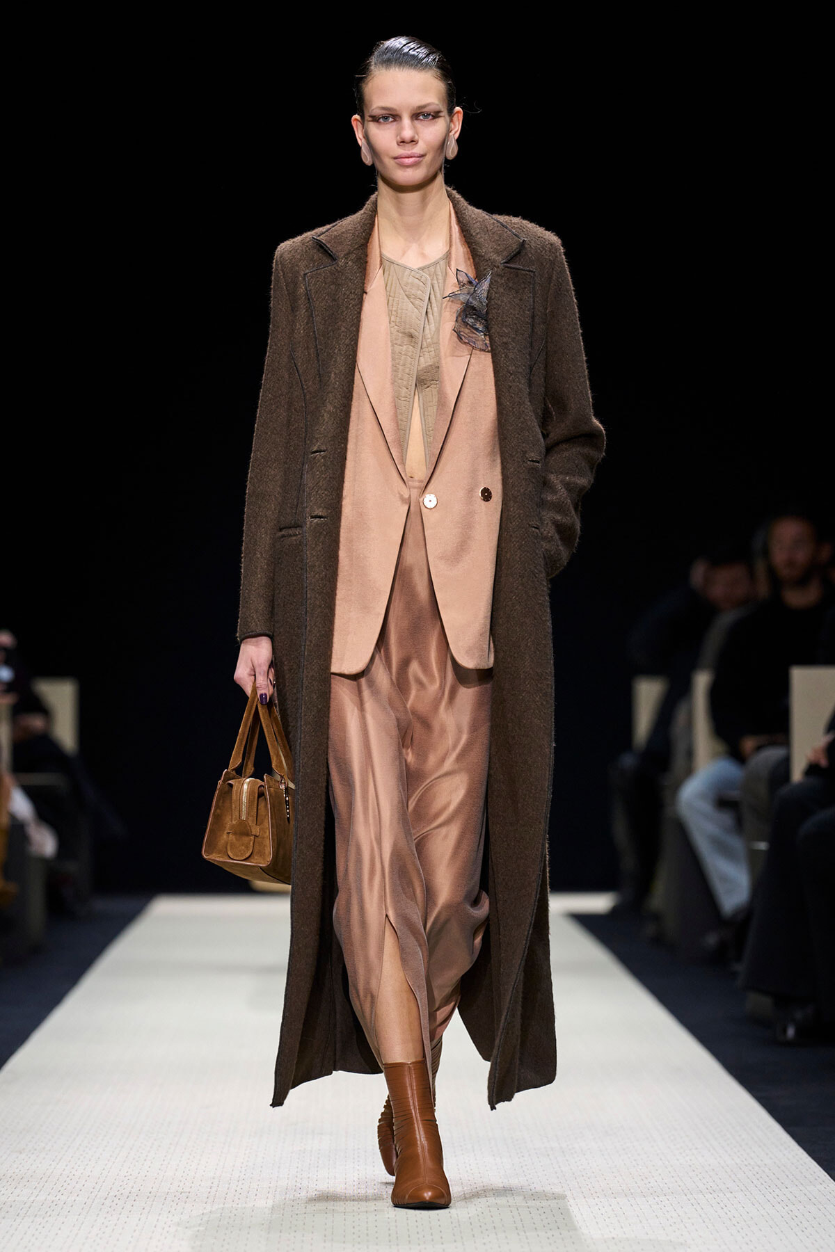 Model walking a runway in a long brown overcoat over a blush-pink suit, carrying a brown handbag and wearing tan boots.