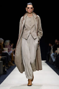 Model walks a fashion runway in a beige layered outfit: long coat with brown piping over a matching vest and light pleated trousers, brown ankle boots, sunglasses.
