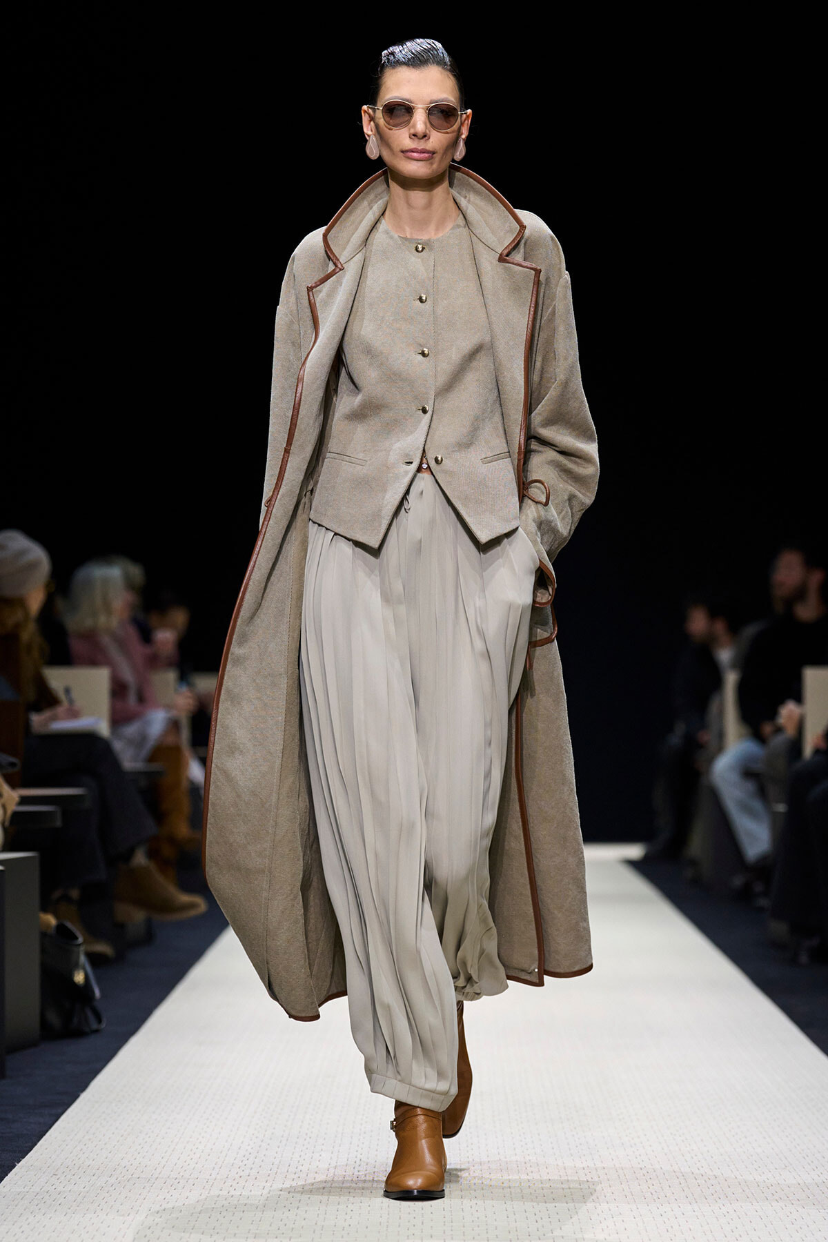 Model walks a fashion runway in a beige layered outfit: long coat with brown piping over a matching vest and light pleated trousers, brown ankle boots, sunglasses.