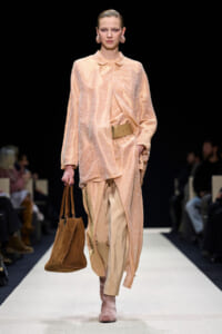 Model walks a fashion runway wearing a peach textured coat over matching pants, cinched with a wide tan belt, carrying a brown suede tote bag.
