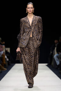 Model walking a fashion runway in a brown and blue patterned suit with matching pants, holding a small brown clutch bag.